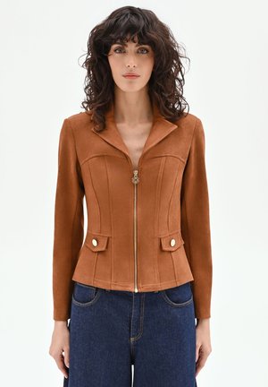 Femme aux cheveux bouclés foncés portant une veste marron ajustée à fermeture éclair avec des boutons dorés et un jean bleu taille haute, debout devant un fond blanc.