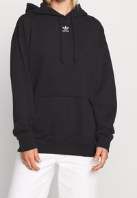 Černá mikina Adidas s velkou přední kapsou, kapucí na šňůrku a minimálním bílým logem na hrudi. Materiál směsi bavlny.