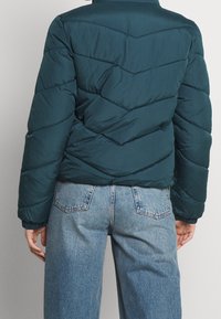 Tyrkisgrøn dunjakke med chevron-quiltning, høj krave og elastiske manchetter, kombineret med lyseblå jeans med vide ben. Tekstureret stoffinish.