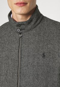 Šedý sako s rybí kostí má texturu, vysoký límec, dvě černá knoflíky, stříbrný zip a malé černé logo na levé straně.