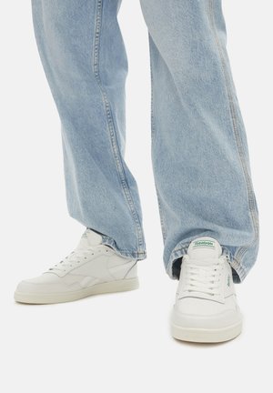 Jeans de cintura alta de color azul claro de pierna ancha, combinados con zapatillas blancas de Reebok con detalles del logo en verde, sobre un fondo liso.