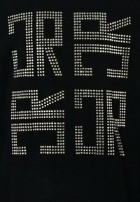 Czarny t-shirt z projektowanym napisem ozdobionym srebrnymi ćwiekami. Napis składa się z wyraźnych, geometrycznych liter utworzonych przez błyszczące ćwieki, tworzących teksturalny efekt wizualny.