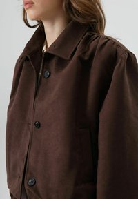 Bruine jas met een zachte textuur en een klassieke kraag, voorzien van grote knopen en zijzakken, met een gestructureerde maar ontspannen uitstraling.