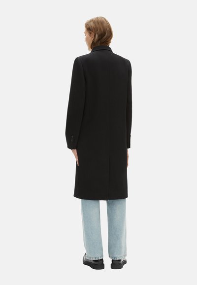 Manteau long noir en tissu texturé avec une coupe droite, doté d'un col classique et de manches longues, associé à un jean large bleu clair.