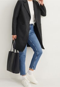 Cappotto grigio in lana con rever a punta, due tasche, indossato su una t-shirt bianca, abbinato a jeans blu e sneakers bianche. Borsa tote nera in mano.