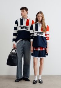 Νεαρός άνδρας και γυναίκα φοράνε ταιριαστά ράγκμπι μπλουζάκια Tommy Hilfiger· ο άνδρας κρατάει μαύρη τσάντα duffle, η γυναίκα φοράει φούστα καρό και μαύρα παπούτσια.