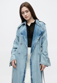 Jovem mulher com cabelo castanho liso usa um longo casaco trench coat de ganga leve sobre uma camisola preta e uma saia branca canelada, em pé contra um fundo branco.