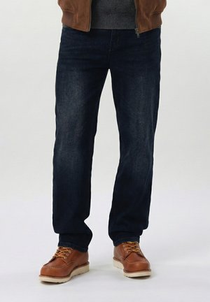 Mann iført mørkeblå jeans og brune skinnstøvler med hvite såler, stående mot en ensfarget lys bakgrunn.