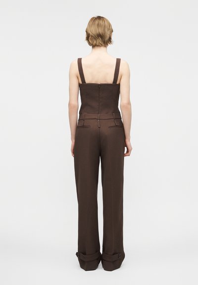 Γυναίκα με κοντά ξανθά μαλλιά που φοράει σκουρό καφέ αμάνικο εφαρμοστό τοπ και ταιριαστό παντελόνι με φαρδιά πόδια και μανσέτες, στεκόμενη με την πλάτη προς το μέρος μας.