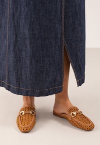 Zuecos marrones trenzados con detalle de hardware dorado, combinados con una falda de mezclilla larga que tiene una abertura lateral y costuras en contraste.