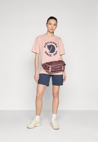 Gaiši rozā T-krekls ar tumšu logo, tumši zils šorti, brūns jostas somiņa un bēšas kedas. Modelis stāv pretī vienkāršai baltai fona.