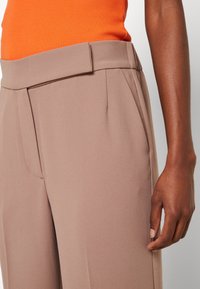 Ljust bruna skräddarsydda byxor med veck, med en slät tygstruktur och en platt midja. En orange ribbad topp syns vid midjan.