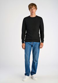 Schwarzer strukturierter Pullover, blaue gerade Jeans und weiße Sneakers. Das Outfit hat einen Rundhalsausschnitt und enganliegende Bündchen am Pullover.