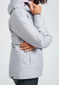 Personne portant une veste à capuche gris clair, une main touchant le menton, l'autre bras croisé, pantalon noir avec l'étiquette ROXY visible.