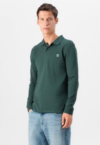 Tummanvihreä pitkähihainen poolopaita, jossa on kaulus, ja rinnassa pieni vaaleanvihreä logo. Paita on valmistettu pehmeästä puuvillasekoitemateriaalista.