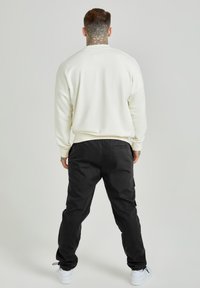 Man med tatueringar på halsen och händerna, iförd vit sweatshirt, svarta byxor och vita sneakers, står vänd bort från kameran mot en grå bakgrund.