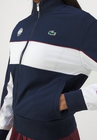 Marinblå zip-up jacka med vita och vinröda horisontella ränder, med en rund logotyp och grön krokodil-emblem på bröstet. Ribbad manschetter.