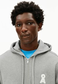 Grijze hoodie met rits, gemaakt van zachte stof, met een ritssluiting aan de voorkant en een wit logo-accent. Draagt over een blauw T-shirt met ronde hals.