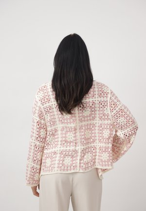 Femme aux longs cheveux noirs portant un cardigan ajouré en crochet rose et blanc et un pantalon clair, vue de dos sur un fond uni.