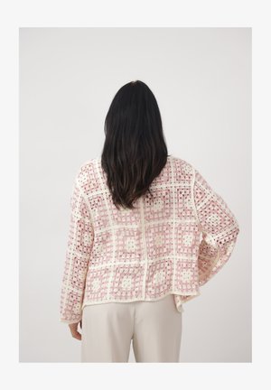 Femme aux longs cheveux noirs portant un cardigan ajouré en crochet rose et blanc et un pantalon clair, vue de dos sur un fond uni.