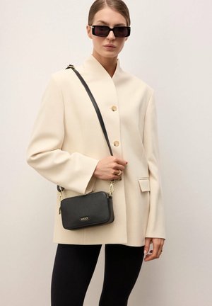 Kobieta w czarnych okularach przeciwsłonecznych, kremowej marynarce, czarnych legginsach, trzymająca czarną torebkę na ramię ze złotymi elementami.