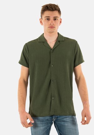 Chemise à manches courtes, col, de couleur vert foncé, en tissu texturé. Présente un devant boutonné et une coupe décontractée. Portée avec un jean bleu.