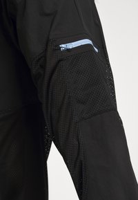 Chaqueta deportiva negra con un panel de tela de malla en las mangas, un bolsillo con cremallera de color azul claro y un material suave en el cuerpo.