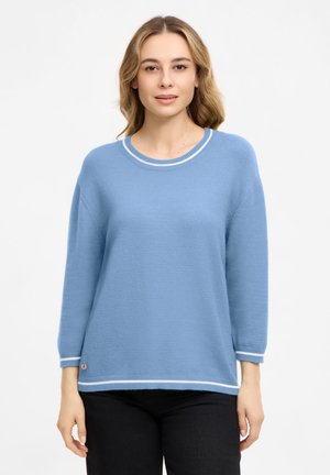 Young woman with light brown hair wearing a light blue long-sleeve sweater with white trim and black pants, standing against a plain background.