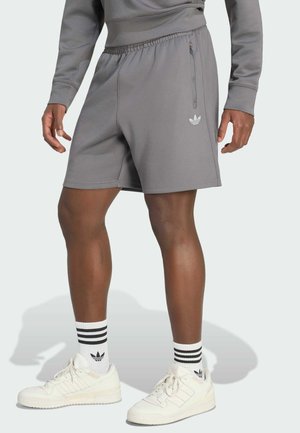 Muž v šedých šortkách Adidas, bílých teniskách Adidas a bílých ponožkách s černými pruhy stojí před jednobarevným pozadím.