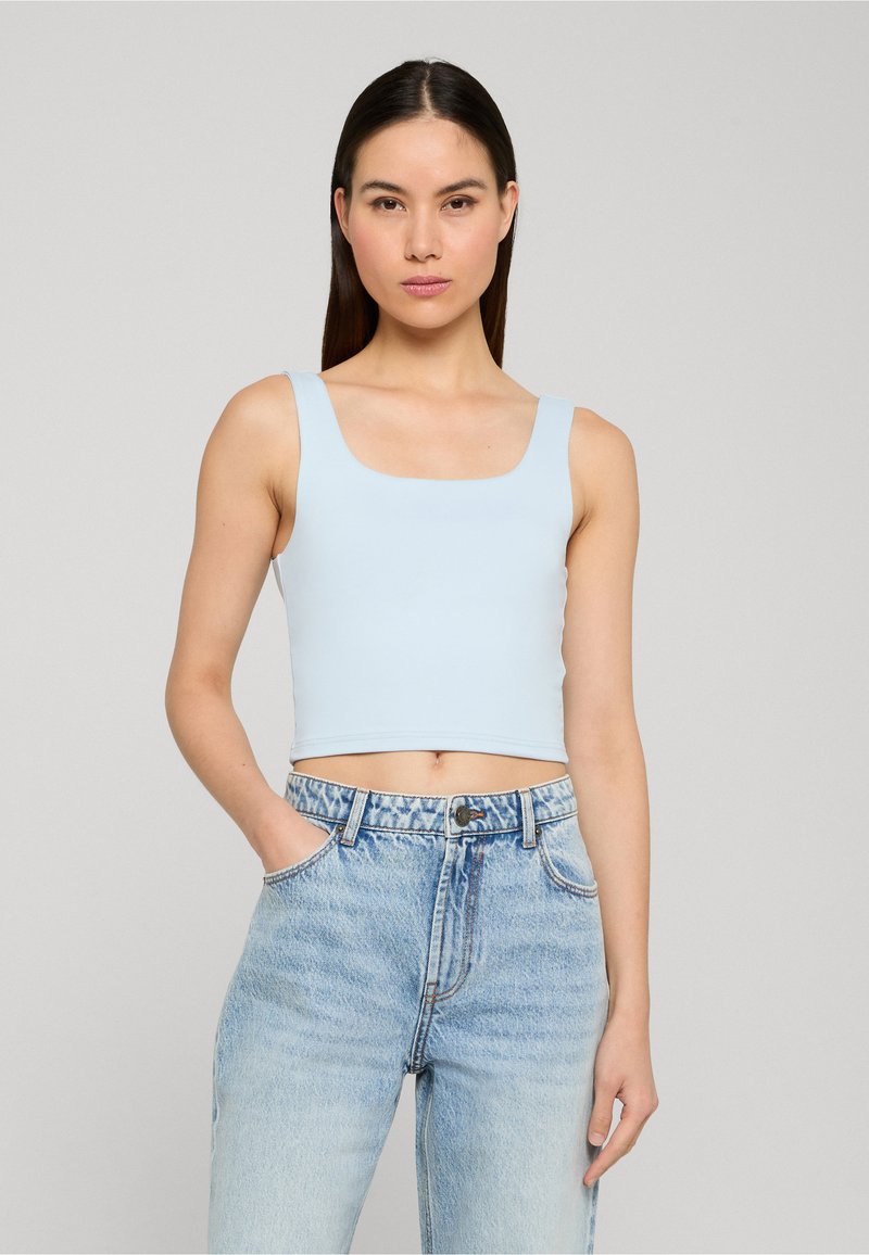 Femme aux cheveux lisses et foncés porte un crop top sans manches bleu clair et un jean taille haute délavé clair sur un fond gris uni.