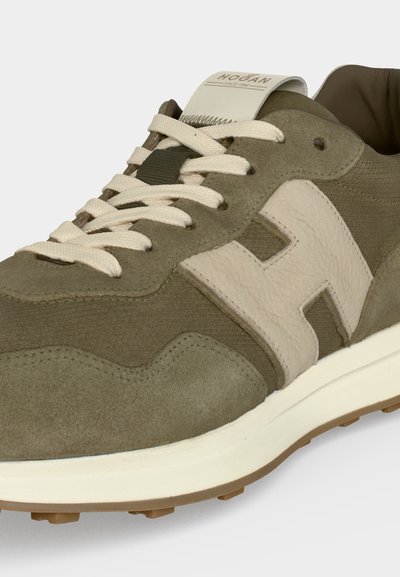 Gros plan d'une basket en daim vert olive et beige avec des lacets blancs, grand logo "H" sur le côté et semelle blanche rembourrée.