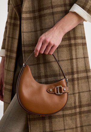 Braune Lederhandtasche mit einer geschwungenen Form, metallischen Hardware-Akzenten und einem seitlichen Tragegurt. Von einer Hand gehalten, vor einem karierten Hintergrund.