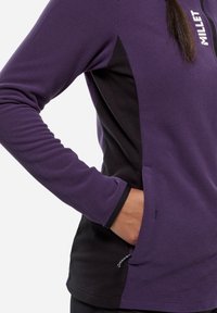 Purple thermal top featuring a textured fabric, black side accents, and zippered pockets. Logo displayed on the chest.