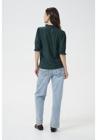 Top verde scuro con maniche a sbuffo e collo arricciato, abbinato a jeans di denim blu chiaro. Il retro presenta un'apertura a chiave e un motivo testurizzato.