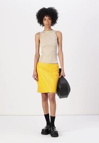 Beige ribbed tanktop en een gele leren kokerrok. Model draagt zwarte platform schoenen en houdt een zwarte tas vast, met knielange zwarte sokken.