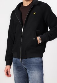 Schwarze Zip-Jacke mit gerippten Bündchen und Saum, aus strukturiertem Stoff gefertigt. Verfügt über ein gesticktes gelbes Logo auf der Brust und seitliche Taschen.