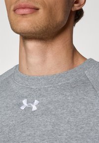 Sweat-shirt gris avec une finition texturée, mettant en avant un col rond et un petit logo blanc sur la poitrine. Gros plan sur les détails du col et du logo.