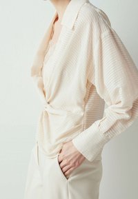 Chemise boutonnée beige clair avec des rayures texturées, grand col et manches longues, nouée à la taille, associée à un pantalon crème.