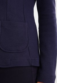 Navy modrá texturovaná bunda s viditelnou kapsou a jemnými švy. Materiál vypadá silně a odolně s pevnějším střihem.