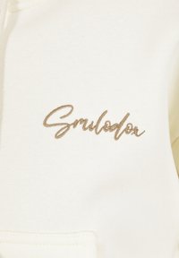 Texte cursif brodé beige « Smilodon » sur un tissu écru, au-dessus d'une couture horizontale ou d'un bord de poche cousu.