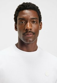 White cotton T-shirt with a crew neck, featuring a small textured logo patch on the left chest. Smooth surface and basic fit design.