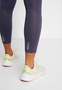 Navy leggings with large perforated design, paired with light-colored athletic shoes featuring a neon green accent and textured fabric.