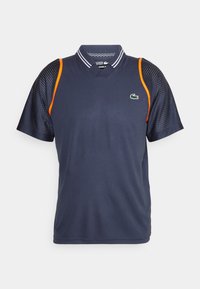 Marinblå sportpolo med nätärmar, orange detaljer och en grön logotyp. Har en klassisk krage och andningsbart tyg.
