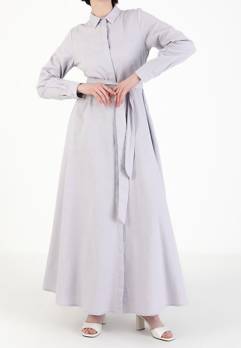 Robe longue grise clair avec un col, un devant à boutons et des manches longues. Comprend une ceinture à nouer à la taille. Tissu au toucher lisse.