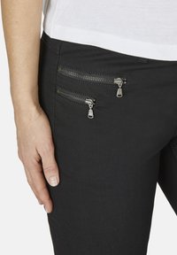 Svarta byxor med två dragkedjefickor på framsidan, prydda med silverfärgad hårdvara. Tyget verkar vara glatt med en figurnära design.