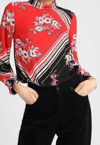 Blusa con motivo floreale rosso e righe nere, colletto alto e maniche lunghe. Abbinata a pantaloni in velluto a coste neri. Tessuto lucido.