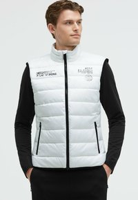 Chaleco acolchado blanco con cuello alto, cierre de cremallera yaccesorios negros. Presenta logotipos y texto en la parte delantera. Lleva puesto sobre una camiseta negra de manga larga.