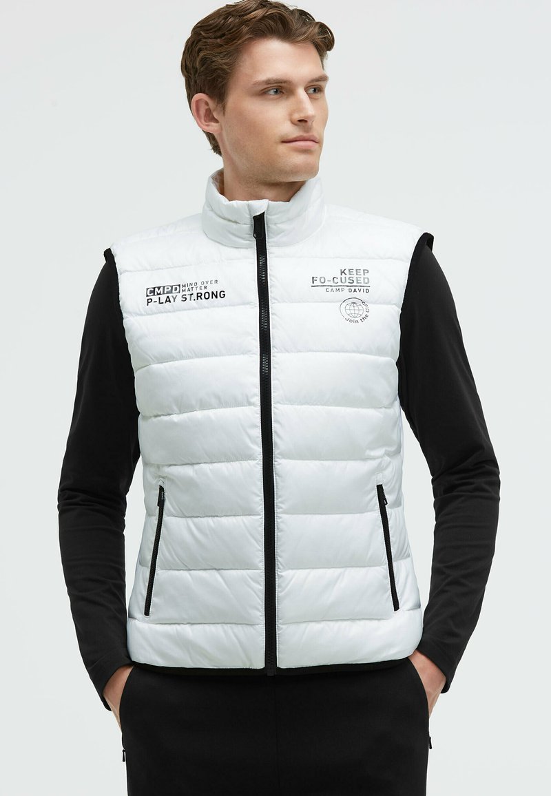 Chaleco acolchado blanco con cuello alto, cierre de cremallera yaccesorios negros. Presenta logotipos y texto en la parte delantera. Lleva puesto sobre una camiseta negra de manga larga.