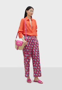Orange skjorta med knappar, mönstrade rosa och blå vida byxor, rosa sandaler, vävd handväska med rosa detaljer och monogram.