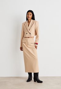 Beige taillierter Blazer mit kontrastierendem Nähen, kombiniert mit einem passenden high-waisted Rock. Schwarze Stiefel mit Absatz runden das Outfit ab.
